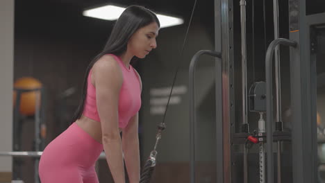A-brunette-woman-in-a-pink-suit-pulls-a-rope-in-a-crossover-arm-for-triceps-training.-Hand-training-in-a-trainer.-Professional-woman-instructor